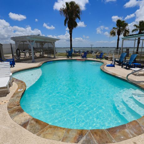 The resort's heated pool, surrounded by scenic views and deck chairs, with guests lounging and kids playing in the water at Copano Bay RV Resort in Rockport, TX.