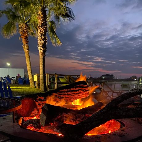 A glowing fire pit at dusk surrounded by palm trees, located at Copano Bay RV Resort in Rockport, TX.