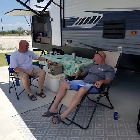 Two men seated in chairs beside an RV at Copano Bay RV Resort in Rockport, TX, enjoying a leisurely moment outdoors.